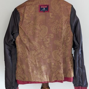 Op de afbeelding: Een rood leren jack met een bruine paisley voering. Het jack heeft een ritssluiting en een opstaande kraag. Het label op de achterkant luidt "IMPER PIEL ORIGINAL LEATHER".