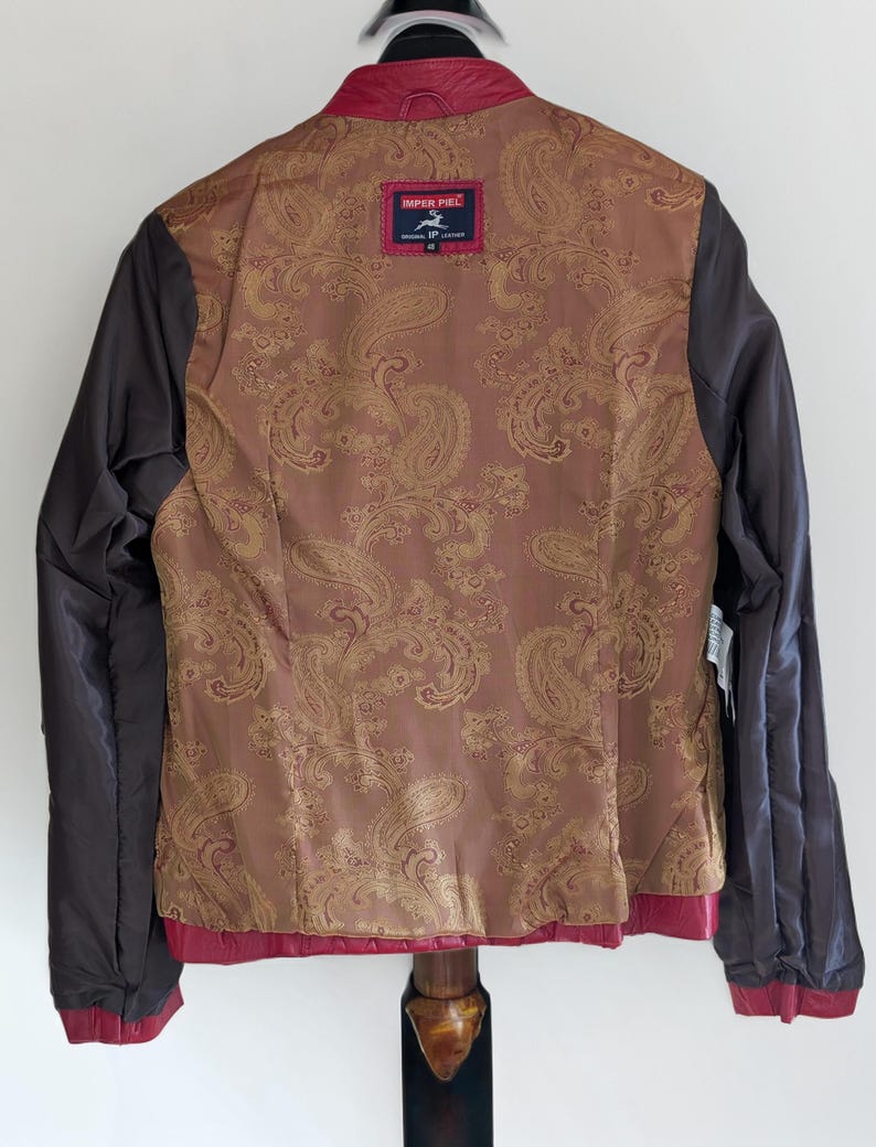 Op de afbeelding: Een rood leren jack met een bruine paisley voering. Het jack heeft een ritssluiting en een opstaande kraag. Het label op de achterkant luidt "IMPER PIEL ORIGINAL LEATHER".