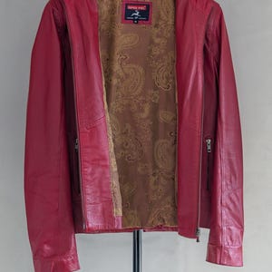 Op de afbeelding: Een rood leren jack met een ritssluiting en een paisley-voering. Het jack heeft twee ritszakken aan de voorkant en een merklabel met de tekst "Imper Piel" met een paardenlogo.
