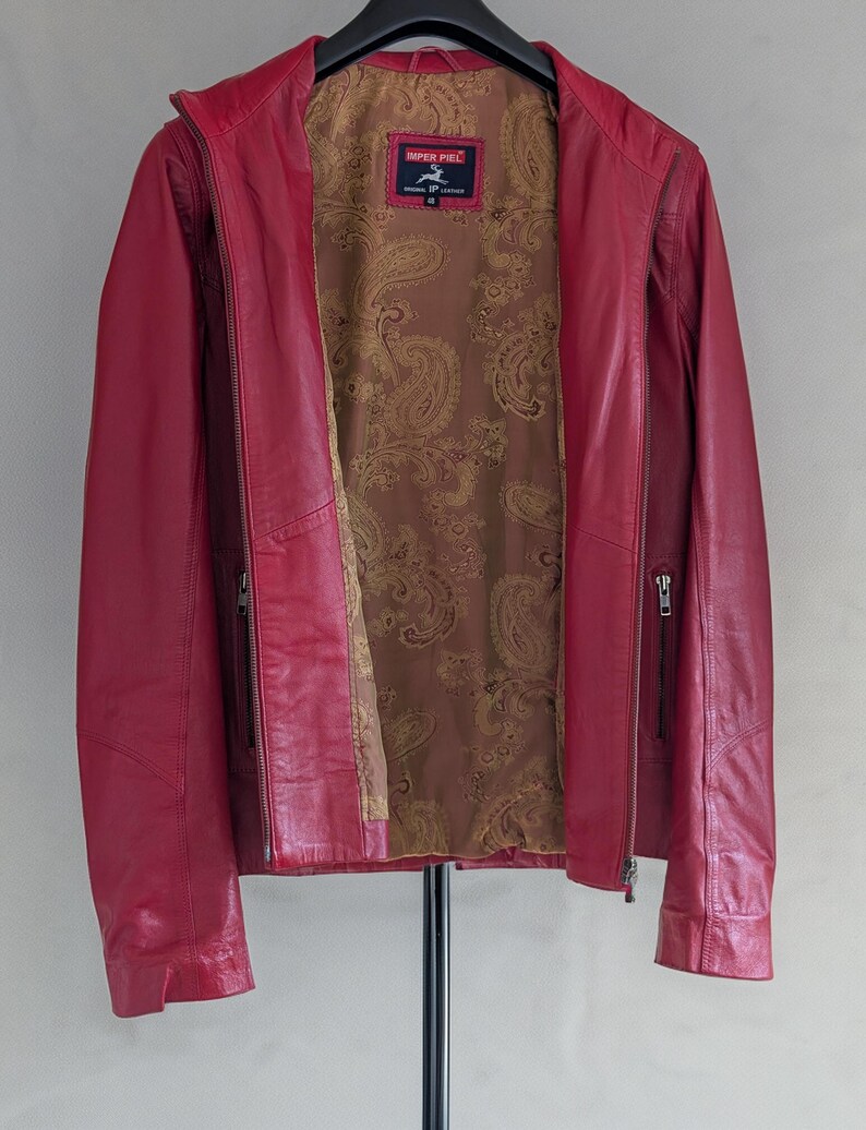 Op de afbeelding: Een rood leren jack met een ritssluiting en een paisley-voering. Het jack heeft twee ritszakken aan de voorkant en een merklabel met de tekst "Imper Piel" met een paardenlogo.