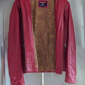 Op de afbeelding: Een rood leren jack met een paisley voering. Het jack heeft een ritssluiting en twee zakken aan de voorkant. Het label op het jack luidt "Imper Piel" en "Echt leer".
