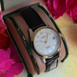 Könnte beinhalten: Eine Timex-Uhr mit silber- und goldfarbenem Gehäuse, hellem Zifferblatt und schwarzem Lederarmband. Das Zifferblatt zeigt die Uhrzeit mit goldfarbenen Ziffern und Zeigern sowie das Datum an. Die Uhr wird in einer Uhrenbox präsentiert.