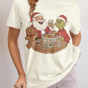 May include: Cream-colored t-shirt featuring a festive illustration of Santa Claus, the Grinch, and a gingerbread man gathered around a table with a bowl of punch. The Grinch and Santa wear red hats and sweaters. The gingerbread man has a bow tie.