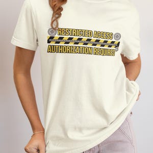 以下が含まれることがあります： 黒と黄色の「RESTRICTED ACCESS AUTHORIZATION REQUIRED」の文字が入ったクリーム色のTシャツ。デザインには、危険テープ風のグラフィックが含まれています。このシャツはカジュアルな半袖スタイルです。