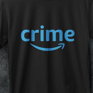 Può includere: T-shirt nero con la parola "crime" in azzurro, sopra una freccia blu curva. La maglietta è appesa a una gruccia bianca. L'immagine include anche un logo per "AA STORE 16 PREMIUM T SHIRT" e una promozione "ACQUISTA 3 GRATIS".