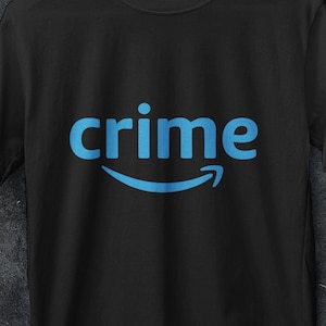 Può includere: T-shirt nero con la parola "crime" in azzurro, sopra una freccia blu curva. La maglietta è appesa a una gruccia bianca. L'immagine include anche un logo per "AA STORE 16 PREMIUM T SHIRT" e una promozione per "ACQUISTA 3 GRATIS".