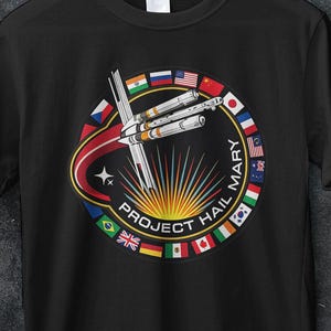 Peut inclure: T-shirt noir avec un motif circulaire représentant un vaisseau spatial, des drapeaux et le texte "PROJECT HAIL MARY". Le design comprend le texte "AA STORE 16 PREMIUM T SHIRT". Le t-shirt contient également du texte promotionnel: "50% OFF MINIMUM BUY 5 CODE: BULK50" et "BUY 3 FREE Code: SHIP3."