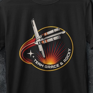 Può includere: T-shirt nera con grafica di un'astronave e la scritta "TEAM GRACE & ROCKY" all'interno di un cerchio dorato. Il design include una scia di cometa rossa e uno sfondo a forma di sole. La maglietta è appesa a una gruccia bianca.