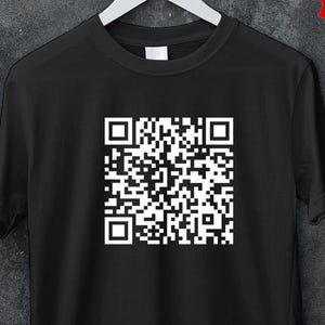 Può includere: T-shirt nero con una grande grafica a codice QR bianca sul davanti. La maglietta è appesa a una gruccia bianca su uno sfondo grigio scuro. Un adesivo promozionale rosso e bianco recita "50% OFF MINIMUM BUY 5 CODE: BULK50". Un adesivo giallo dice "BUY 3 FREE Code: SHIP3".
