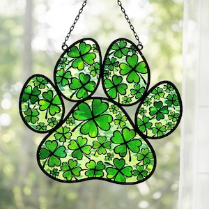 May include: A stained glass paw print decoration with a black outline and a chain for hanging. The paw print is filled with green shamrocks on a light green background, perfect for St. Patrick's Day.
