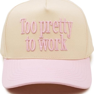 May include: A cream-colored baseball cap with a pink brim and the embroidered text "Too pretty to work" in pink lettering on the front panel. The cap features a structured crown with a button on top and stitched detailing on the brim.