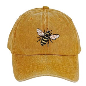 Peut inclure: Casquette de baseball jaune moutarde avec une broderie de motif d'abeille. L'abeille est noire et jaune avec des ailes blanches. La casquette a une visière incurvée et un bouton sur le dessus.