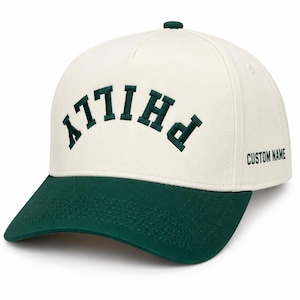 Puede incluir: Una gorra de béisbol color crema con visera verde oscuro y un botón verde oscuro en la parte superior. El frente presenta la palabra PHILLY bordada en una fuente arqueada verde oscuro. El lateral de la gorra incluye el texto CUSTOM NAME.
