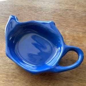 May include: A cobalt blue ceramic sauce dish with a handle. The dish has a unique, organic shape with a slightly raised rim. The handle is a simple loop, and the dish sits on a wooden surface.