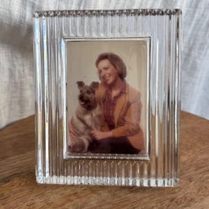 May include: A rectangular clear glass picture frame with a vertical ribbed texture. The frame holds a photograph of a person with light brown hair wearing a tan blazer and a plaid shirt, sitting next to a small, light-colored terrier dog.