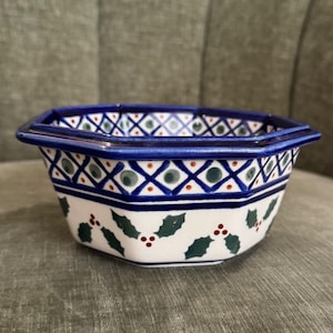 May include: An octagonal ceramic bowl featuring a white base with a hand-painted pattern of dark green holly leaves and small red berries. The rim is decorated with a blue lattice design containing green dots and small red accents.