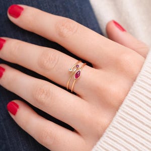 Peut inclure: Trois bagues en or avec des pierres précieuses. L'une est ornée d'un petit diamant, une autre d'un rubis rond et la troisième d'un rubis en forme de marquise. Les bagues sont portées sur une main avec du vernis à ongles rouge.