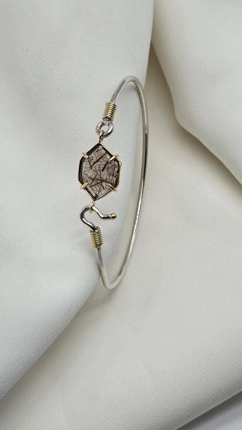 Pu&ograve; includere: Un bracciale in argento con un ciondolo a forma di gemma esagonale. Il ciondolo &egrave; incorniciato in oro e presenta inclusioni scure. Il bracciale ha un design curvo con dettagli dorati.