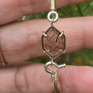 Pu&ograve; includere: Un braccialetto fatto a mano con una gemma esagonale marrone con inclusioni interne, incastonata in una lunetta color oro. Il braccialetto ha un cinturino color argento con accenti di filo color oro e un elemento di design curvo decorativo.