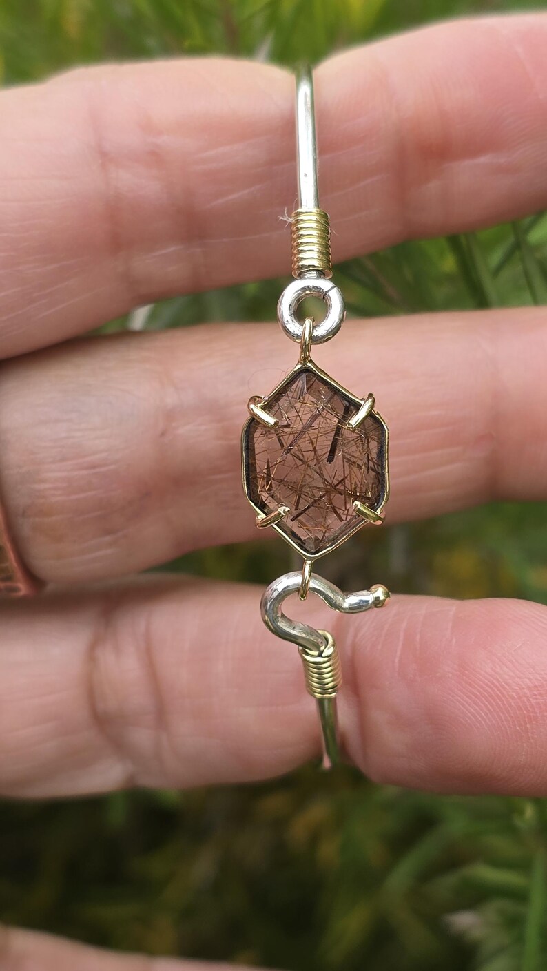 Pu&ograve; includere: Un braccialetto fatto a mano con una gemma esagonale marrone con inclusioni interne, incastonata in una lunetta color oro. Il braccialetto ha un cinturino color argento con accenti di filo color oro e un elemento di design curvo decorativo.