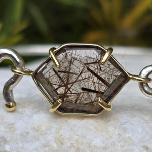 Pu&ograve; includere: Bracciale con pietra preziosa esagonale con centro trasparente e inclusioni marroni, incastonata in una lunetta color oro. Il bracciale ha maglie curve color argento su ogni lato. La gemma &egrave; lunga circa 2,5 cm.