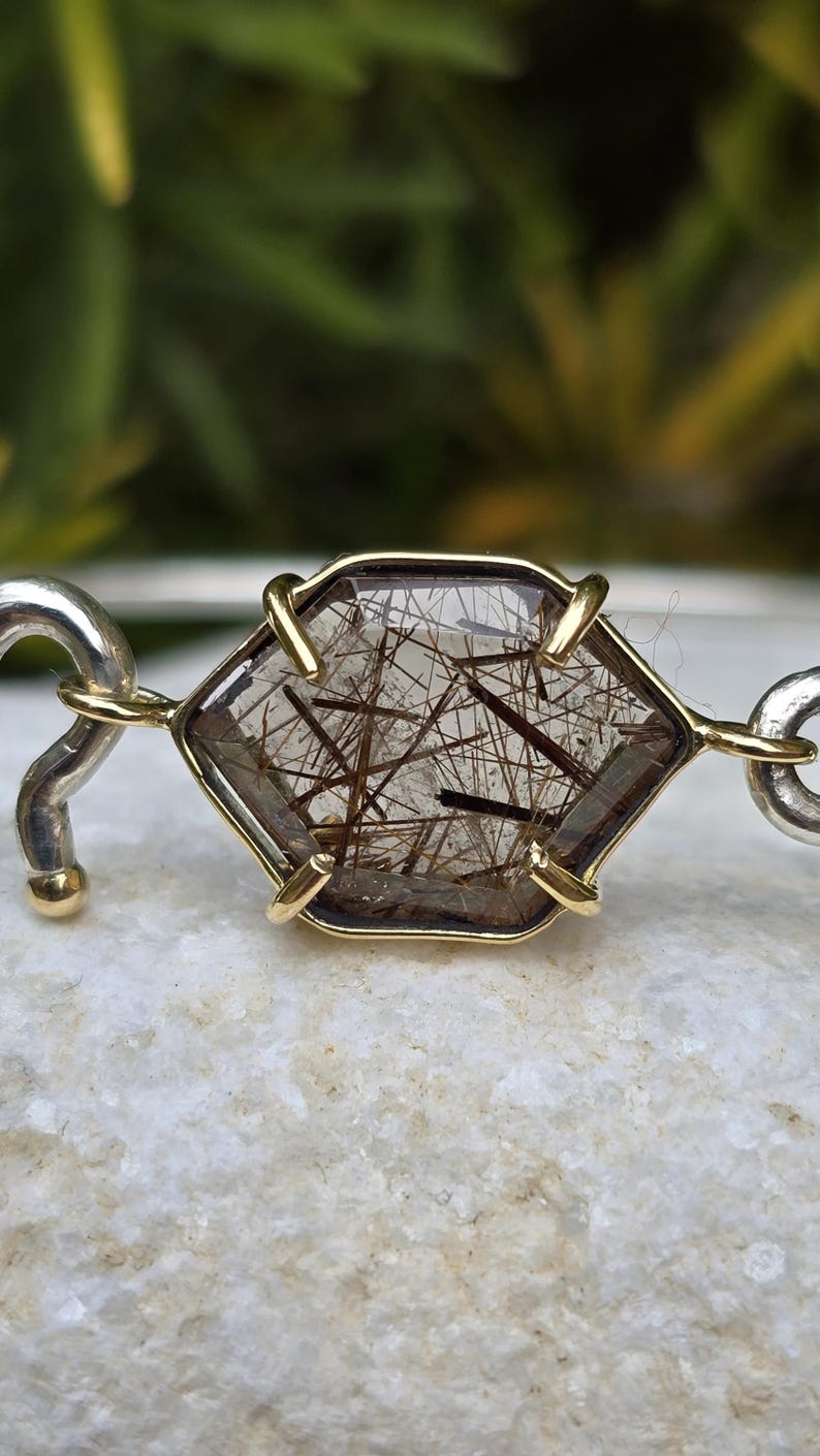 Pu&ograve; includere: Bracciale con pietra preziosa esagonale con centro trasparente e inclusioni marroni, incastonata in una lunetta color oro. Il bracciale ha maglie curve color argento su ogni lato. La gemma &egrave; lunga circa 2,5 cm.