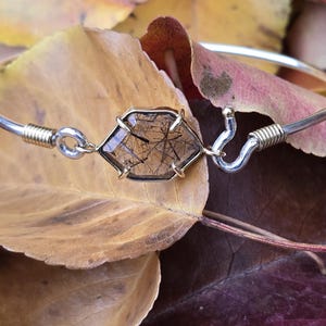 Pu&ograve; includere: Un bracciale rigido argentato con una gemma esagonale trasparente con inclusioni scure. La gemma &egrave; incastonata in una lunetta color oro. Il bracciale &egrave; appoggiato su foglie autunnali, mostrando un'estetica naturale e terrosa.