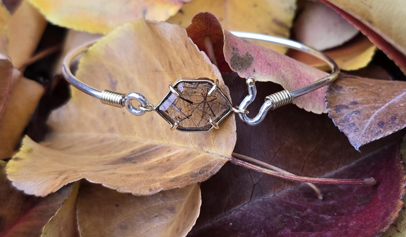 Pu&ograve; includere: Un bracciale rigido argentato con una gemma esagonale trasparente con inclusioni scure. La gemma &egrave; incastonata in una lunetta color oro. Il bracciale &egrave; appoggiato su foglie autunnali, mostrando un'estetica naturale e terrosa.
