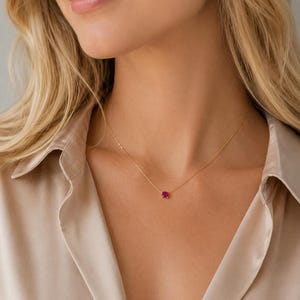 Può includere: Una delicata collana a catena dorata con un singolo piccolo ciondolo in pietra preziosa rosa brillante dal taglio quadrato. La collana poggia sulla clavicola, mostrando un design essenziale con una sottile catena in metallo lucido.
