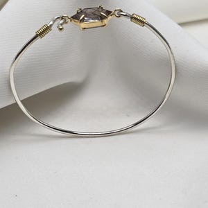 Pu&ograve; includere: Un braccialetto a cerchio d'argento con un elemento centrale in oro e pietra preziosa. La pietra preziosa ha una forma esagonale con una lunetta dorata. Il braccialetto ha dettagli in filo d'oro e una chiusura a gancio. Sfondo bianco.