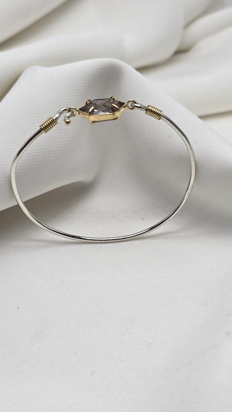 Pu&ograve; includere: Un braccialetto a cerchio d'argento con un elemento centrale in oro e pietra preziosa. La pietra preziosa ha una forma esagonale con una lunetta dorata. Il braccialetto ha dettagli in filo d'oro e una chiusura a gancio. Sfondo bianco.