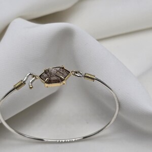 Pu&ograve; includere: Un braccialetto a cerchio in argento con un elemento centrale a forma di gemma esagonale marrone e oro. Il braccialetto ha dettagli dorati ed &egrave; posto su uno sfondo bianco strutturato.