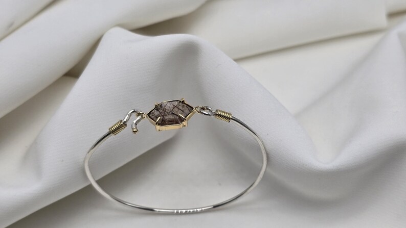 Pu&ograve; includere: Un braccialetto a cerchio in argento con un elemento centrale a forma di gemma esagonale marrone e oro. Il braccialetto ha dettagli dorati ed &egrave; posto su uno sfondo bianco strutturato.