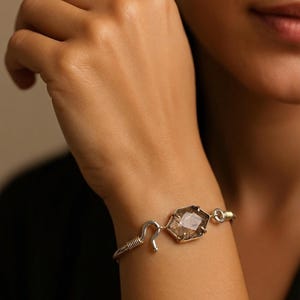 Pu&ograve; includere: Un bracciale in argento con una gemma geometrica, sfaccettata, marrone chiaro. Il bracciale ha una chiusura a gancio e un design delicato avvolto in filo. La gemma &egrave; incastonata in una lunetta color oro.