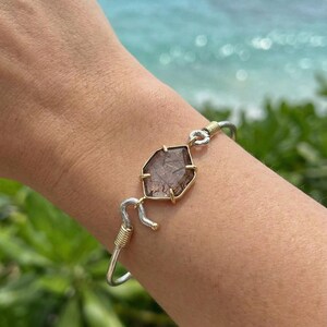 Pu&ograve; includere: Bracciale color argento e oro con una gemma marrone sfaccettata. La gemma &egrave; incastonata in una lunetta dorata e collegata al bracciale d'argento con filo dorato. Il bracciale &egrave; indossato su un polso, con uno sfondo sfocato dell'oceano.