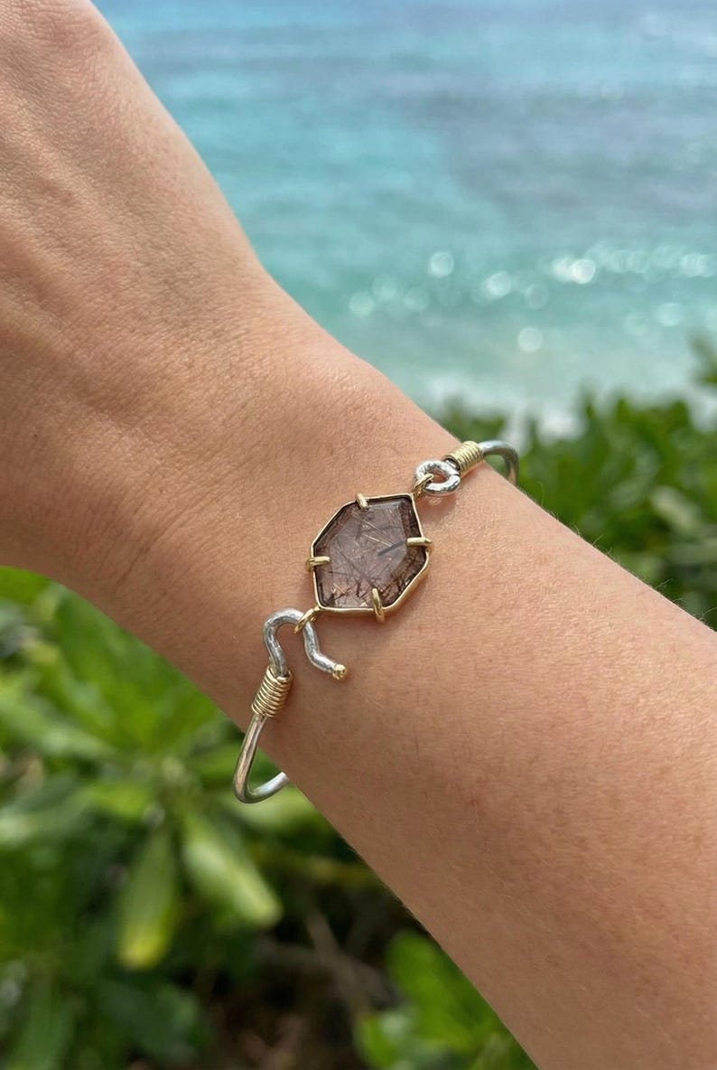 Pu&ograve; includere: Bracciale color argento e oro con una gemma marrone sfaccettata. La gemma &egrave; incastonata in una lunetta dorata e collegata al bracciale d'argento con filo dorato. Il bracciale &egrave; indossato su un polso, con uno sfondo sfocato dell'oceano.