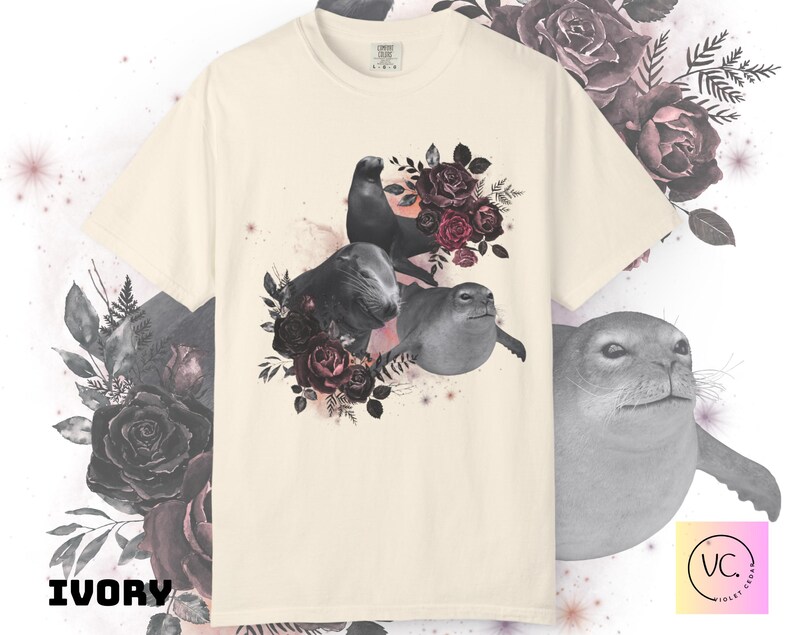 May include: Ivory-colored t-shirt featuring a graphic of three seals surrounded by dark red roses and foliage. The seals are in shades of gray, with one seal perched atop the others. The text "IVORY" is printed in a bold, black font at the bottom left.