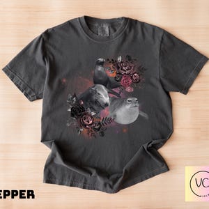 May include: Pepper-colored t-shirt featuring a grayscale illustration of three seals surrounded by a floral arrangement of dark roses and foliage. The design includes a cosmic background. The text "PEPPER" is at the bottom.
