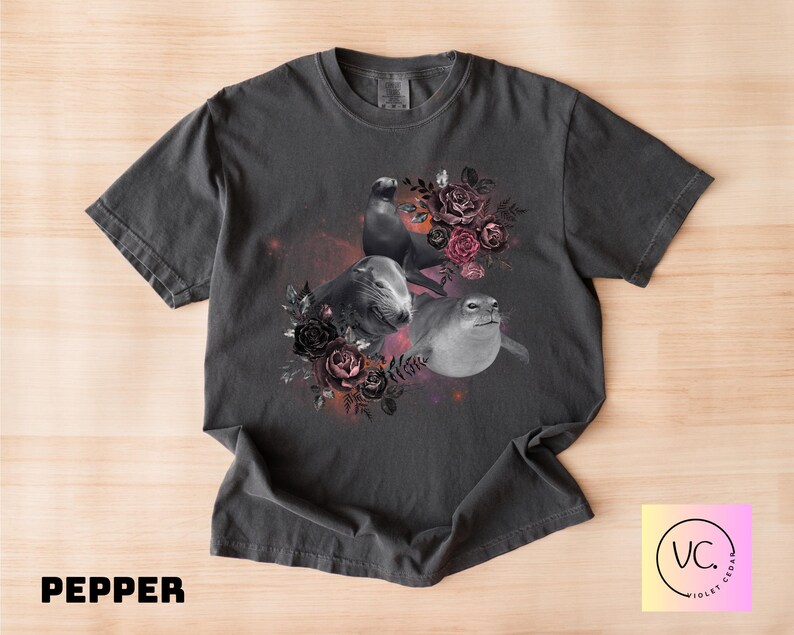 May include: Pepper-colored t-shirt featuring a grayscale illustration of three seals surrounded by a floral arrangement of dark roses and foliage. The design includes a cosmic background. The text "PEPPER" is at the bottom.
