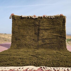 May include: A large, olive green rug with a shaggy texture. The rug has a rectangular shape with a fringe along the bottom edge. The rug is displayed outdoors against a backdrop of a desert landscape and a clear blue sky.