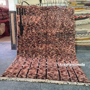 May include: A handmade rug with a geometric pattern in shades of brown and black. The rug has a fringe along the bottom edge and the text "Areahandmade" in white. The rug is displayed on a gray floor.