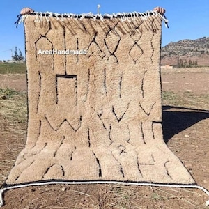 May include: A rectangular beige rug with a thick, shaggy pile and dark brown geometric line patterns. The rug features white fringe at the top and bottom edges, with the text AreaHandmade printed in white on the upper left side.