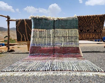 May include: A colorful, rectangular rug with a striped pattern in shades of blue, white, and red. The rug has a fringed edge and is displayed outdoors with other rugs hanging in the background. The rug is made of a thick, woven material.