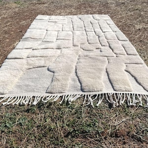 May include: An off-white rectangular rug featuring a raised, textured design of irregular shapes. The rug has a fringe along the bottom edge. The rug is placed on a brown and green surface.