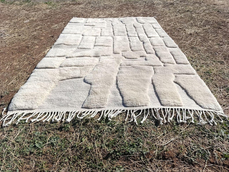 May include: An off-white rectangular rug featuring a raised, textured design of irregular shapes. The rug has a fringe along the bottom edge. The rug is placed on a brown and green surface.