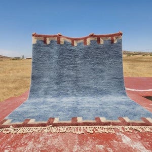 May include: A rectangular area rug featuring a solid blue center with a thick, textured pile. The top and bottom edges are decorated with a repeating geometric pattern in dark red and cream, finished with a fringe of cream-colored tassels.