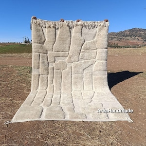 May include: A beige, rectangular rug with a geometric pattern of raised, rectangular shapes. The rug has a fringed edge at the top and is displayed outdoors against a backdrop of a field and a clear blue sky. The text "AreaHandmade" is visible.