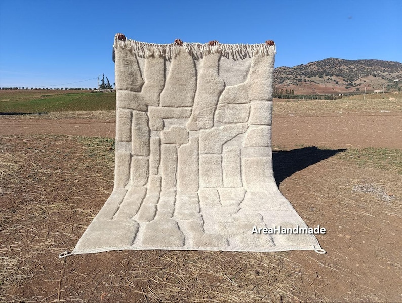 May include: A beige, rectangular rug with a geometric pattern of raised, rectangular shapes. The rug has a fringed edge at the top and is displayed outdoors against a backdrop of a field and a clear blue sky. The text "AreaHandmade" is visible.