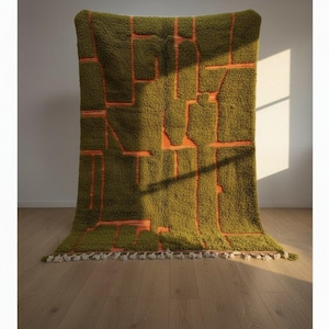 May include: A large, olive green rug with an abstract design in orange. The rug has a shaggy texture and a geometric pattern. The edges are finished with a fringe of white tassels. The rug is displayed against a white wall and wooden floor.