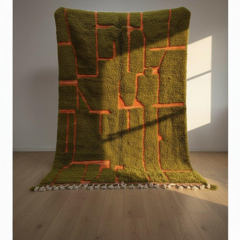 May include: A large, olive green rug with an abstract design in orange. The rug has a shaggy texture and a geometric pattern. The edges are finished with a fringe of white tassels. The rug is displayed against a white wall and wooden floor.